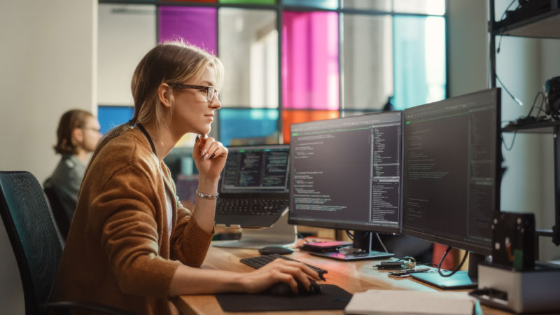 Female Software Engineer Writing Code on Desktop Computer with Multiple Screens Setup in Coworking Office Space. Professional Caucasian Woman Working on SaaS Platform For Innovative Startup